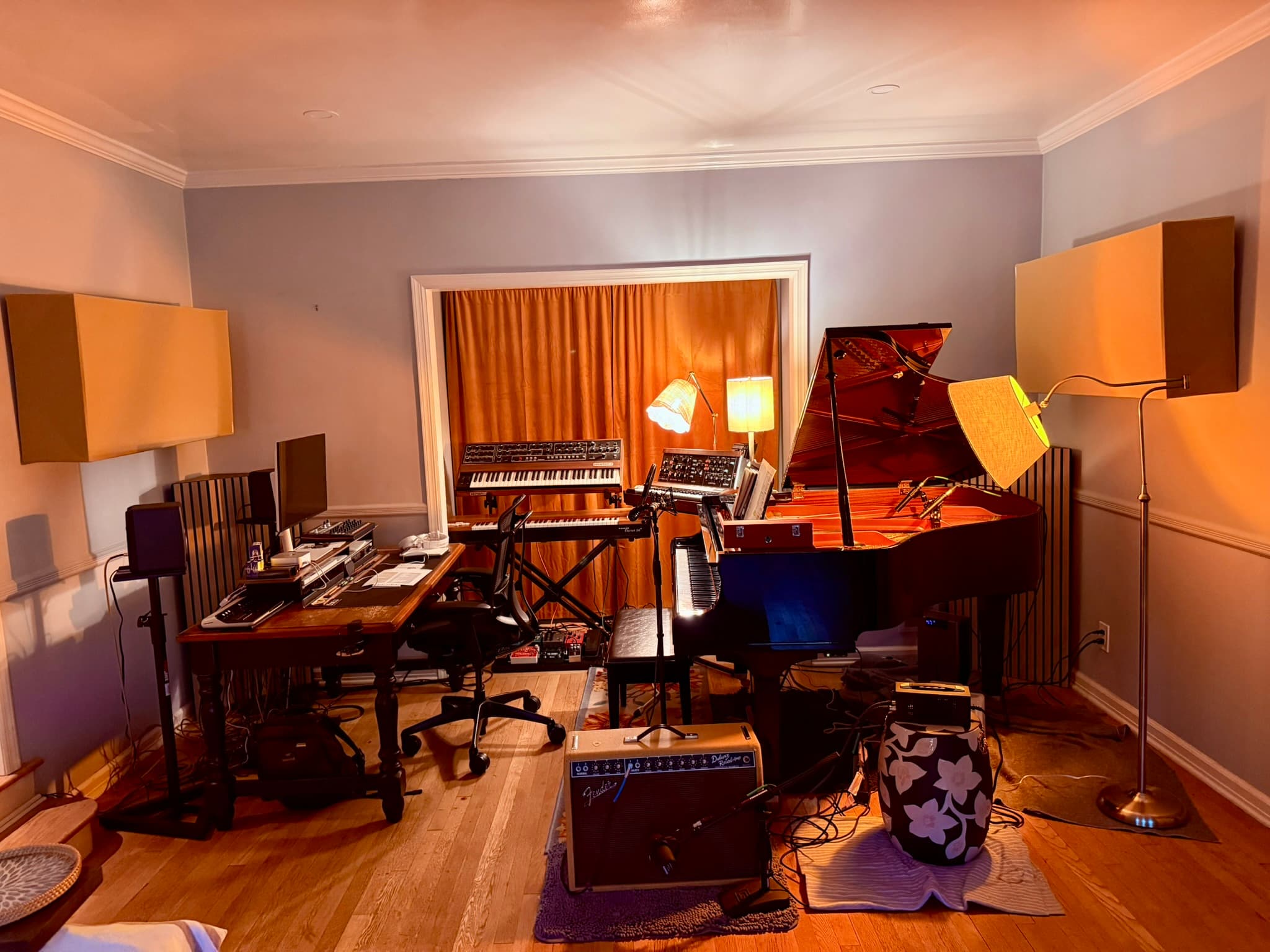 Wide view of the studio — GC1 baby grand on the right, the keys corner and desk on the left, the Deluxe Reverb on the floor, orange curtains in the alcove, GIK SlatFusors in the corners
